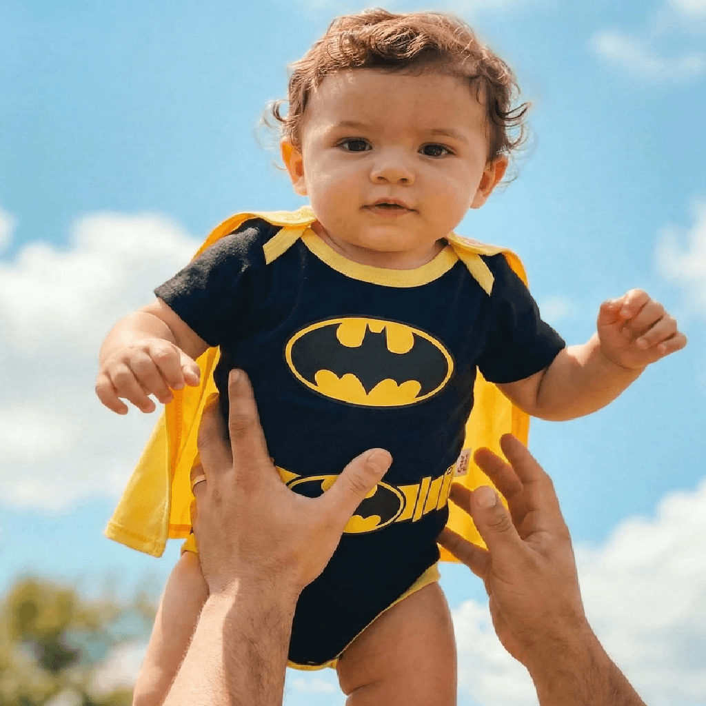 A baby wearing a Batman-themed short-sleeve bodysuit with a yellow cape and matching belt, held up by an adult's hands against a blue sky with clouds.