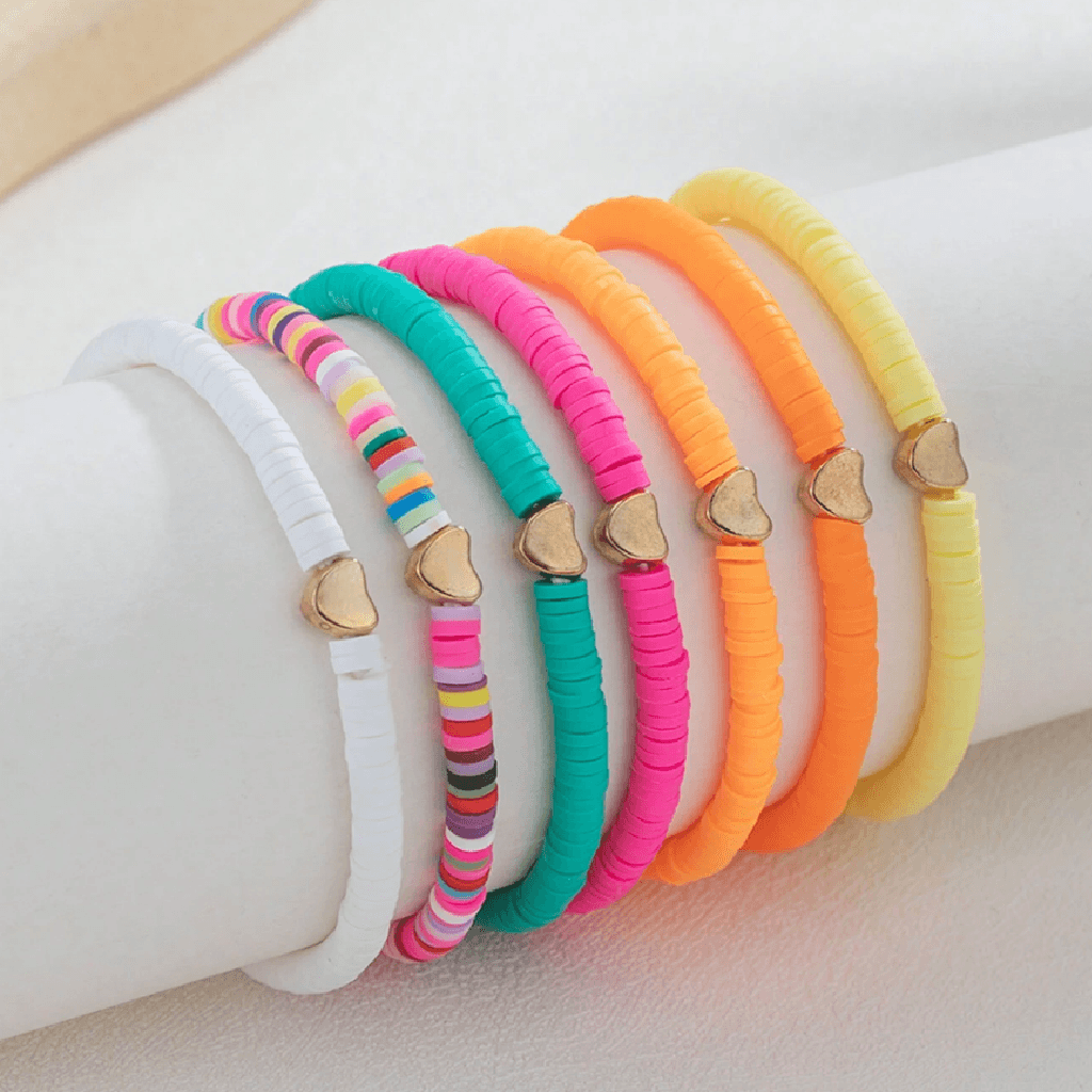 Six colorful braided bracelet bands displayed on a white cylindrical stand. The bands are white, multicolor striped, turquoise, hot pink, neon orange, and pale yellow, each featuring a small gold heart charm.