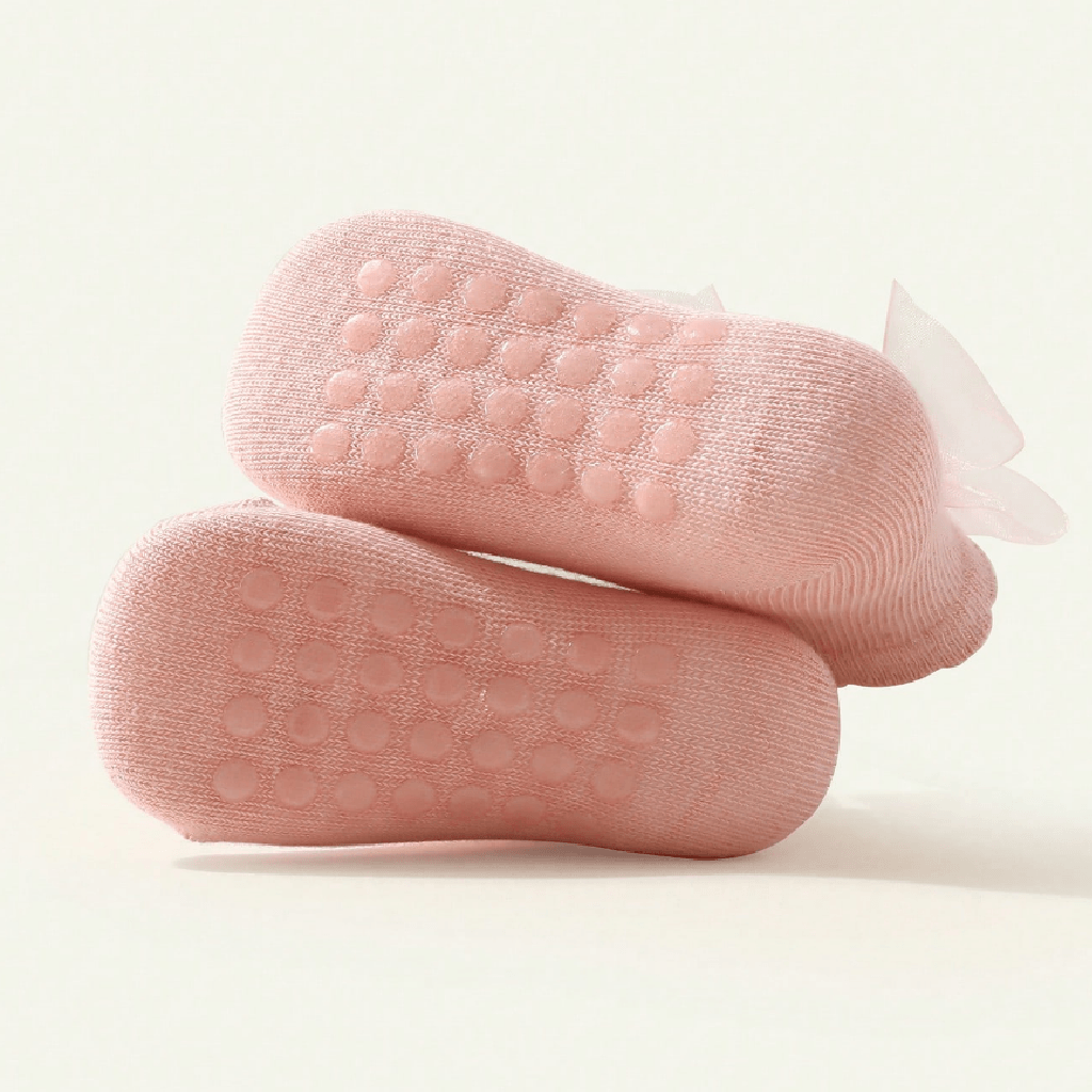 A pair of light pink baby socks with textured non-slip grip dots on the soles and a sheer white ribbon bow on the sides, displayed on a clean off-white background.