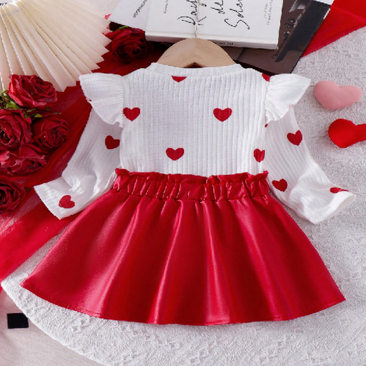 White top with red heart patterns and a red skirt on a textured surface with flowers and books.