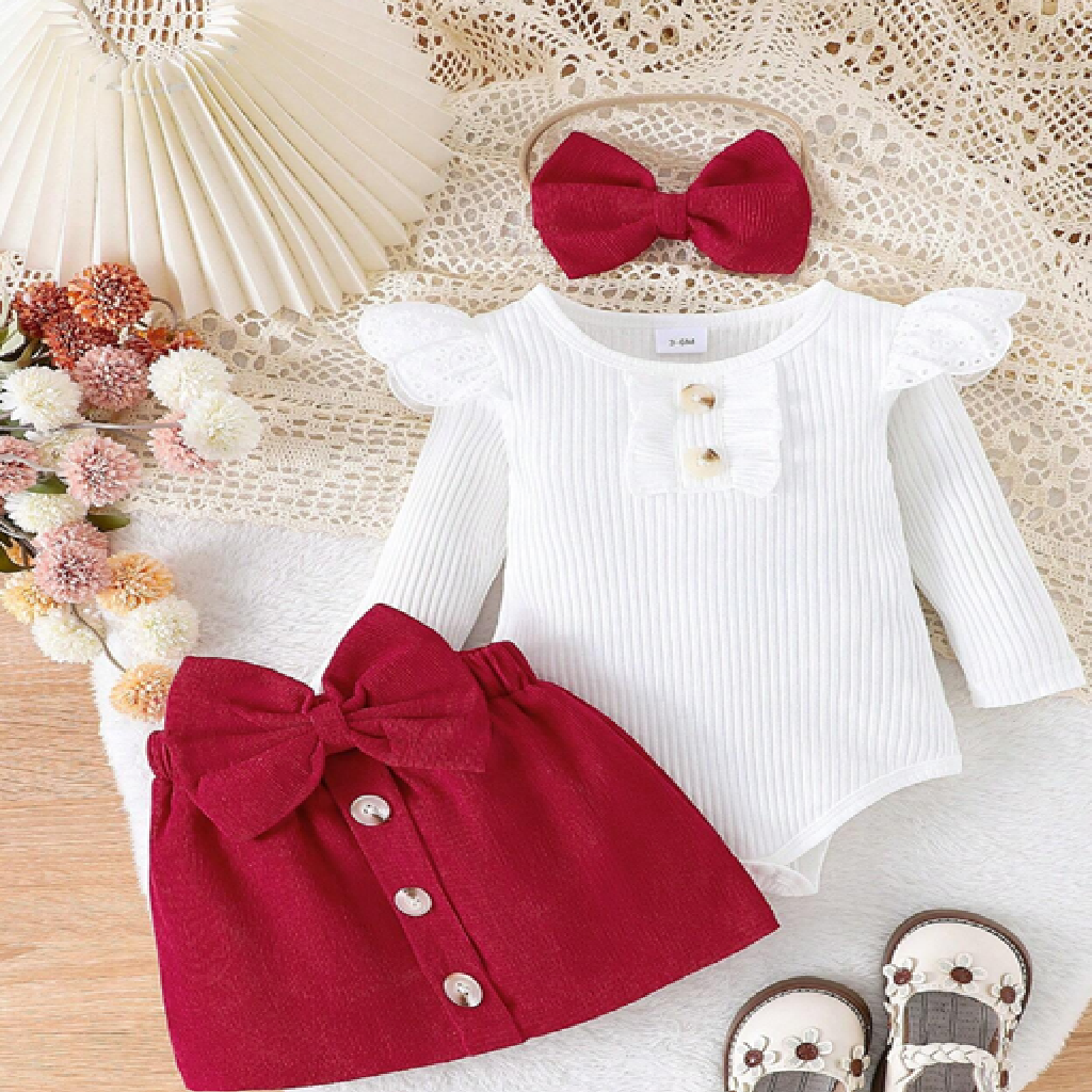 Baby outfit with white long-sleeve top and red skirt on a textured surface with flowers and a hat.
