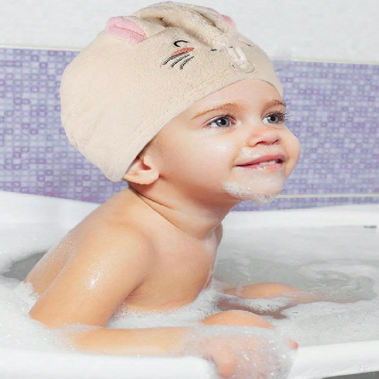 Child in a bathtub wearing a beige towel cap with cartoon face, smiling.