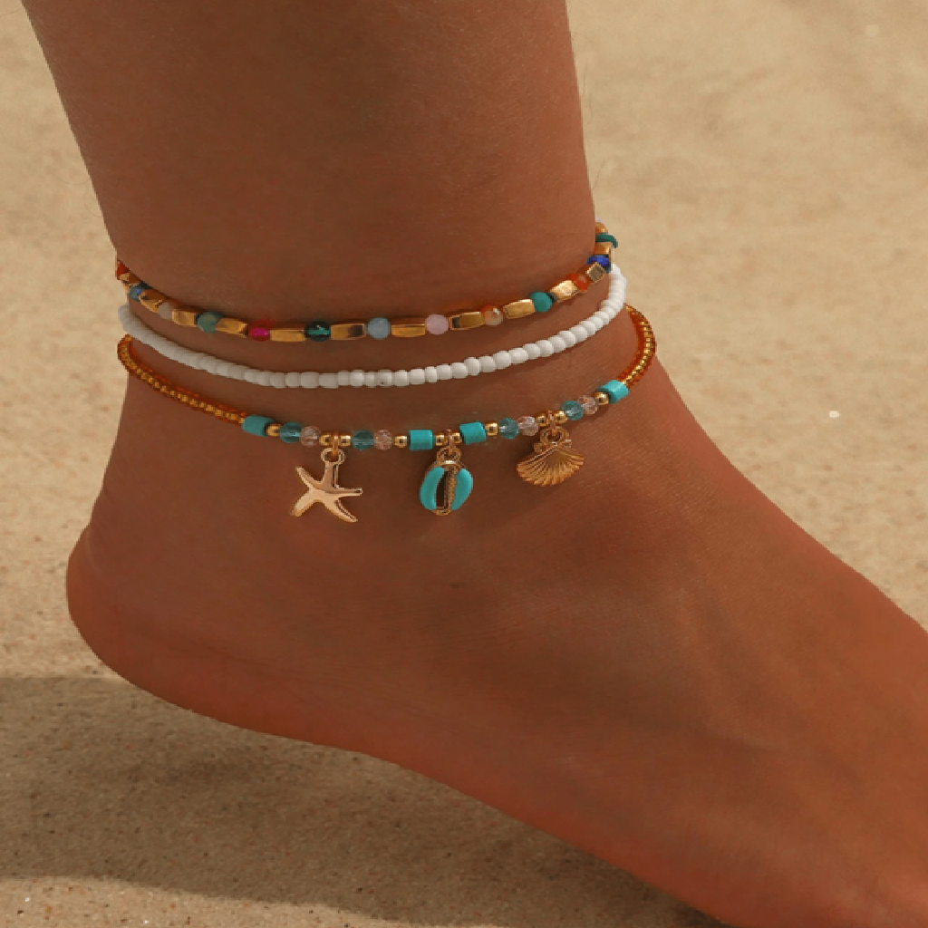 Anchored foot wearing colorful beaded anklets with starfish and shell charms on a sandy background