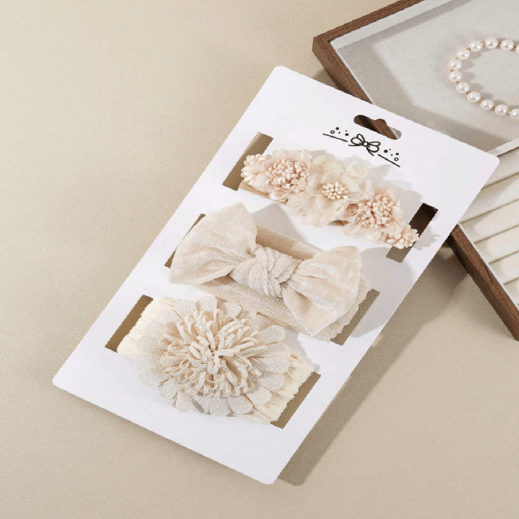 Three cream-colored hair accessories on a white display card, including two fabric bows and one textured flower piece, photographed on a beige surface with a jewelry box partially visible in the background.