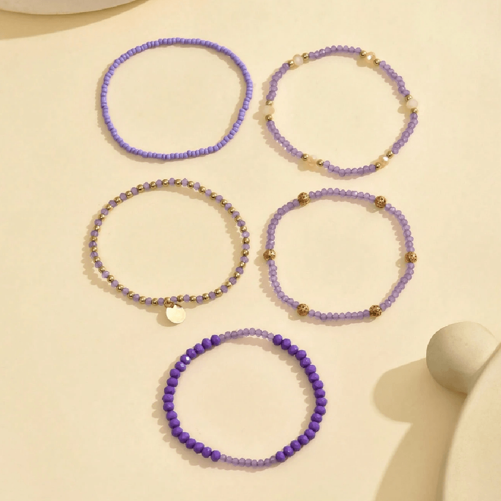Five beaded stretch bracelets arranged on a light cream surface. Four are lavender purple with small gold spacer beads, and one is a deeper violet purple with larger beads and a small round gold charm.
