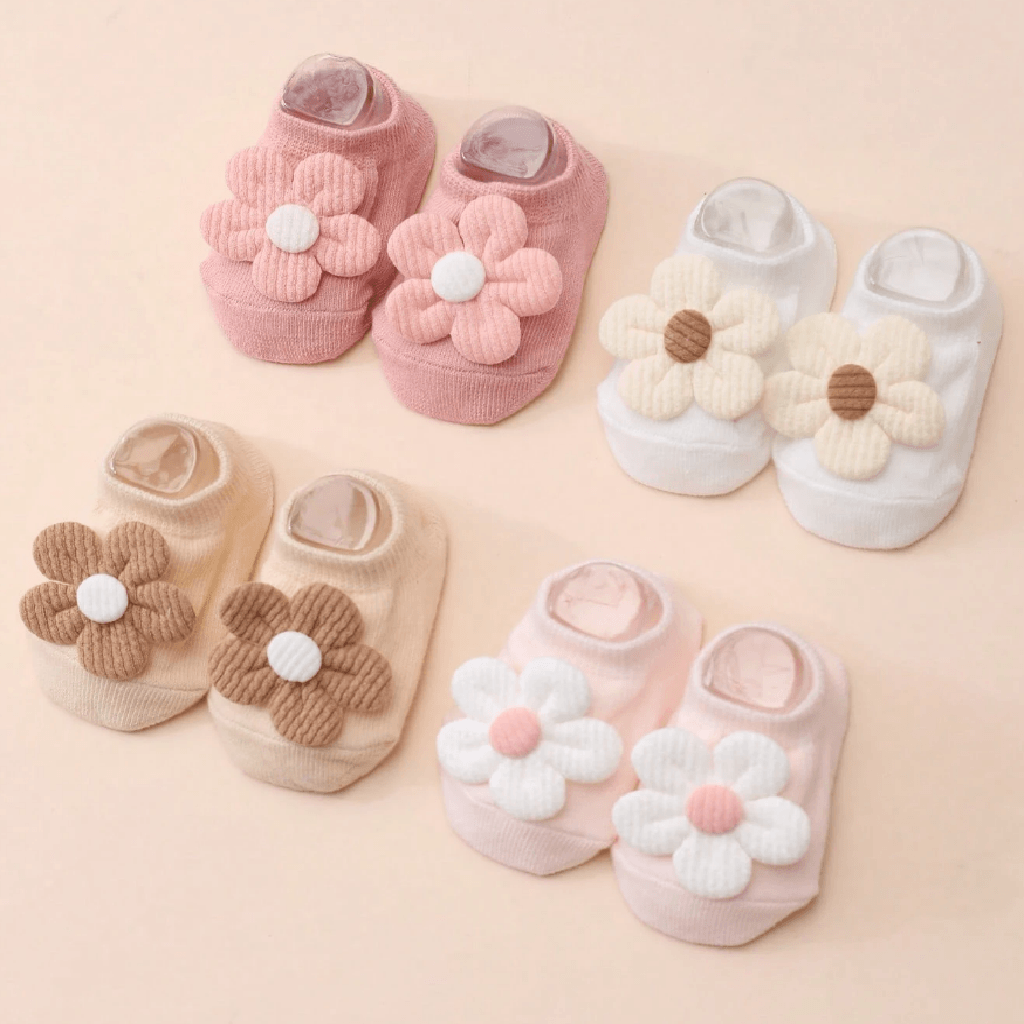 Four pairs of baby slip-on socks arranged on a soft peach background. Each pair features a large flower applique on the front: one pink flower with a white center, one cream flower with a brown center, one tan flower with a white center, and one pale pink flower with a white center.