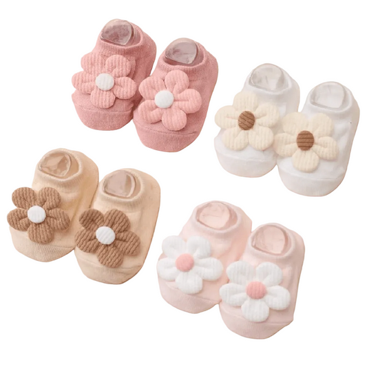 Baby slip-on socks arranged on a soft peach background. Each pair features a large flower appliqué on the front: one pink flower with a white center, one cream flower with a brown center, one tan flower with a white center, and one pale pink flower with a white center.