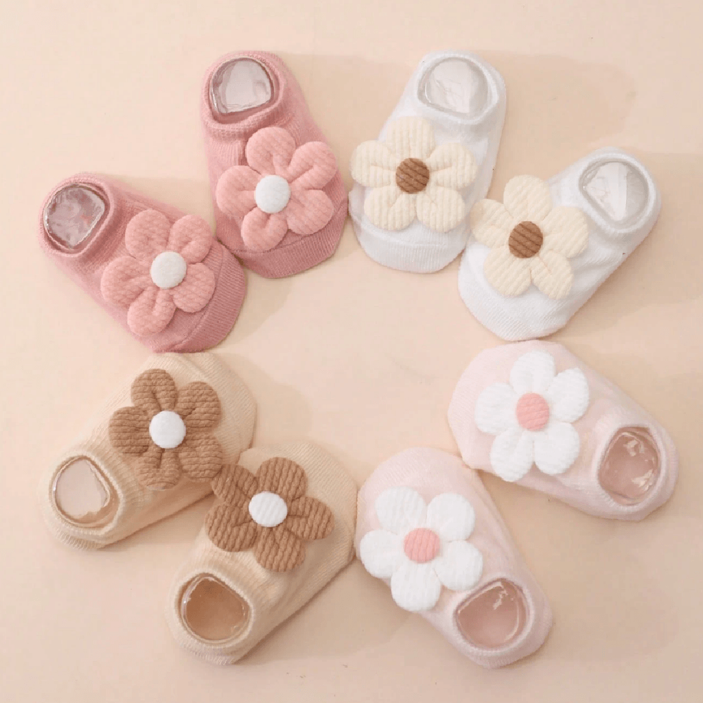 Six pairs of baby slip-on socks arranged in a circle on a soft peach background. Each pair features a large flower applique on the front, with the sock body in pink, white, beige, and light brown tones.