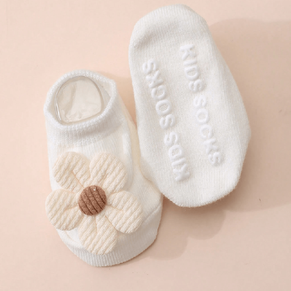 A pair of white knit baby socks on a soft peach background. One sock is shown from the side with a cream-colored rope flower applique and a brown center, and the other is shown from the sole with the words 'KIDS SOCKS' embossed in white.