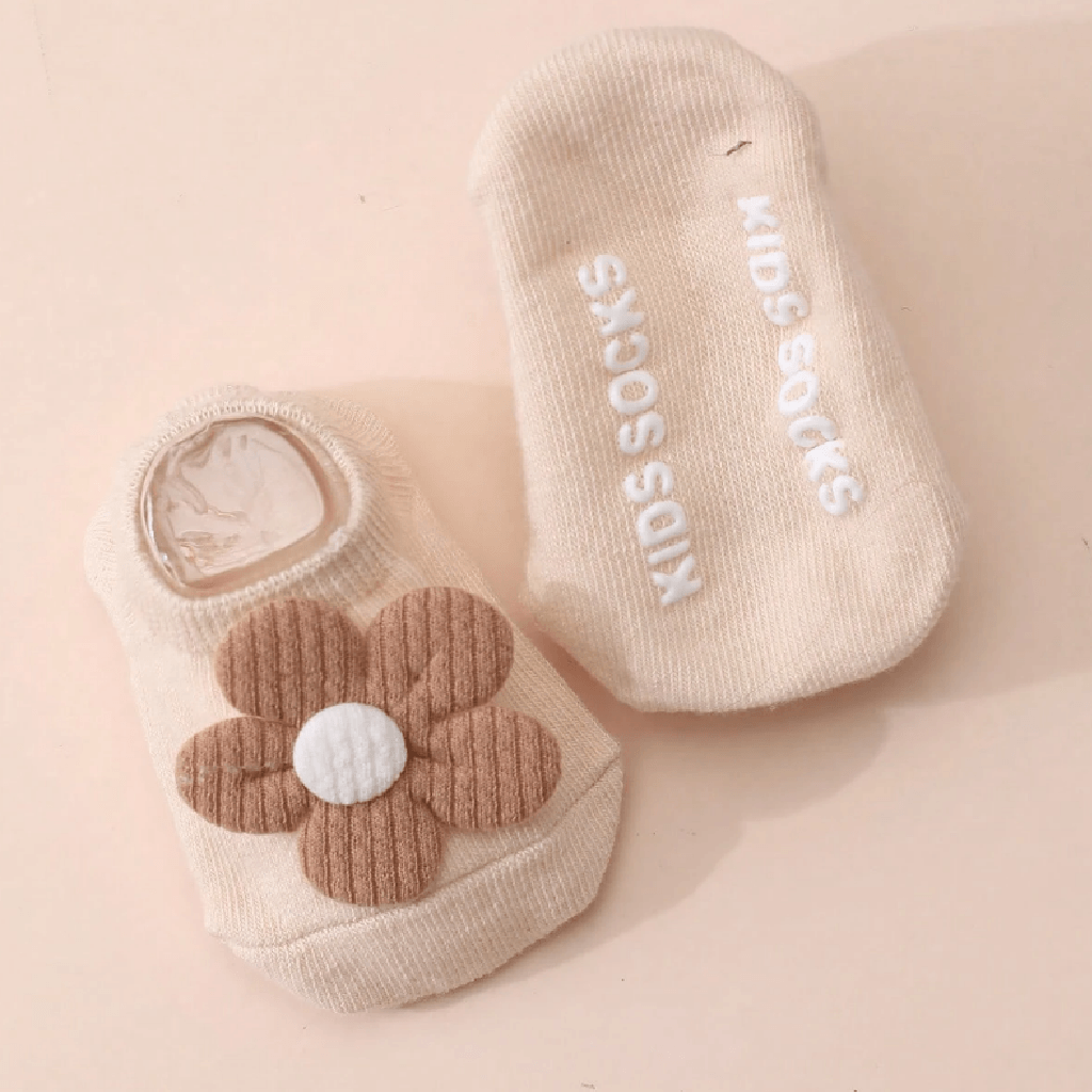 A pair of beige baby socks on a light peach background. One sock features a brown and white corduroy flower applique, while the other displays the white text 'KIDS SOCKS' printed vertically on the sole.