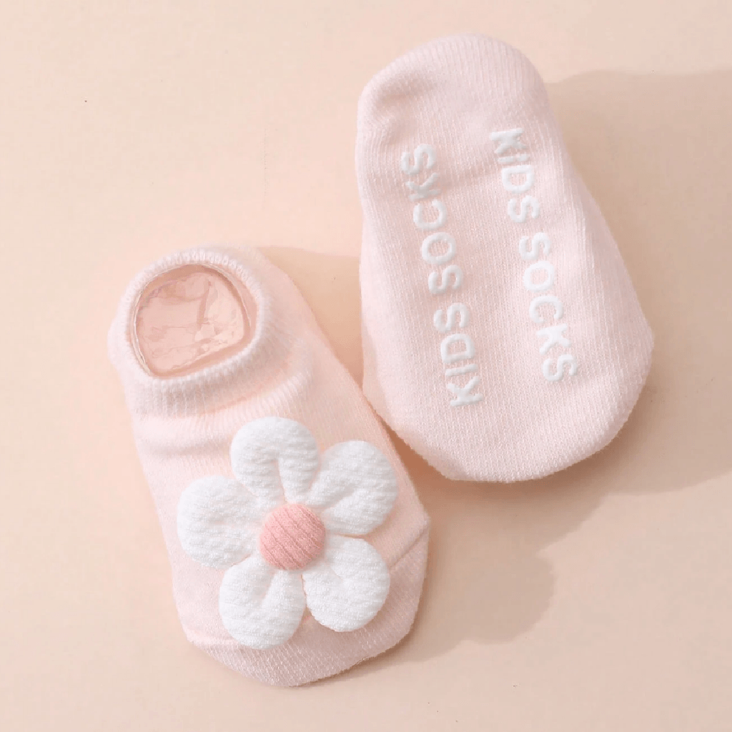 A pair of light pink baby socks on a matching beige background. One sock features a white 3D flower applique with a pink center, and the other sock has 'KIDS SOCKS' printed vertically in white.