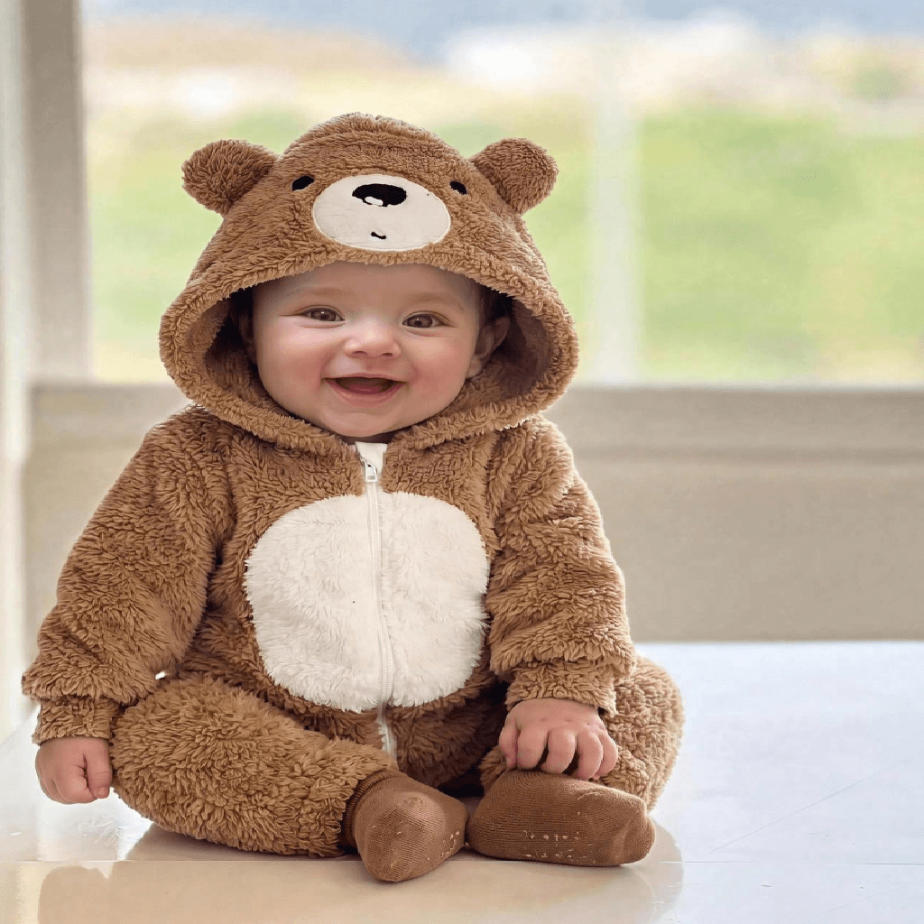 A baby wearing a plush brown bear onesie with a white belly patch and bear face hood, sitting on a light floor in front of a softly blurred window.