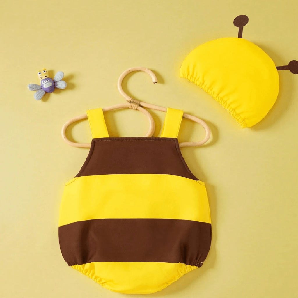 A baby bee-themed two-piece outfit set featuring a yellow and brown striped romper with white flower details on the straps, displayed on a wooden hanger, alongside a matching yellow bee cap on a solid pale yellow background.