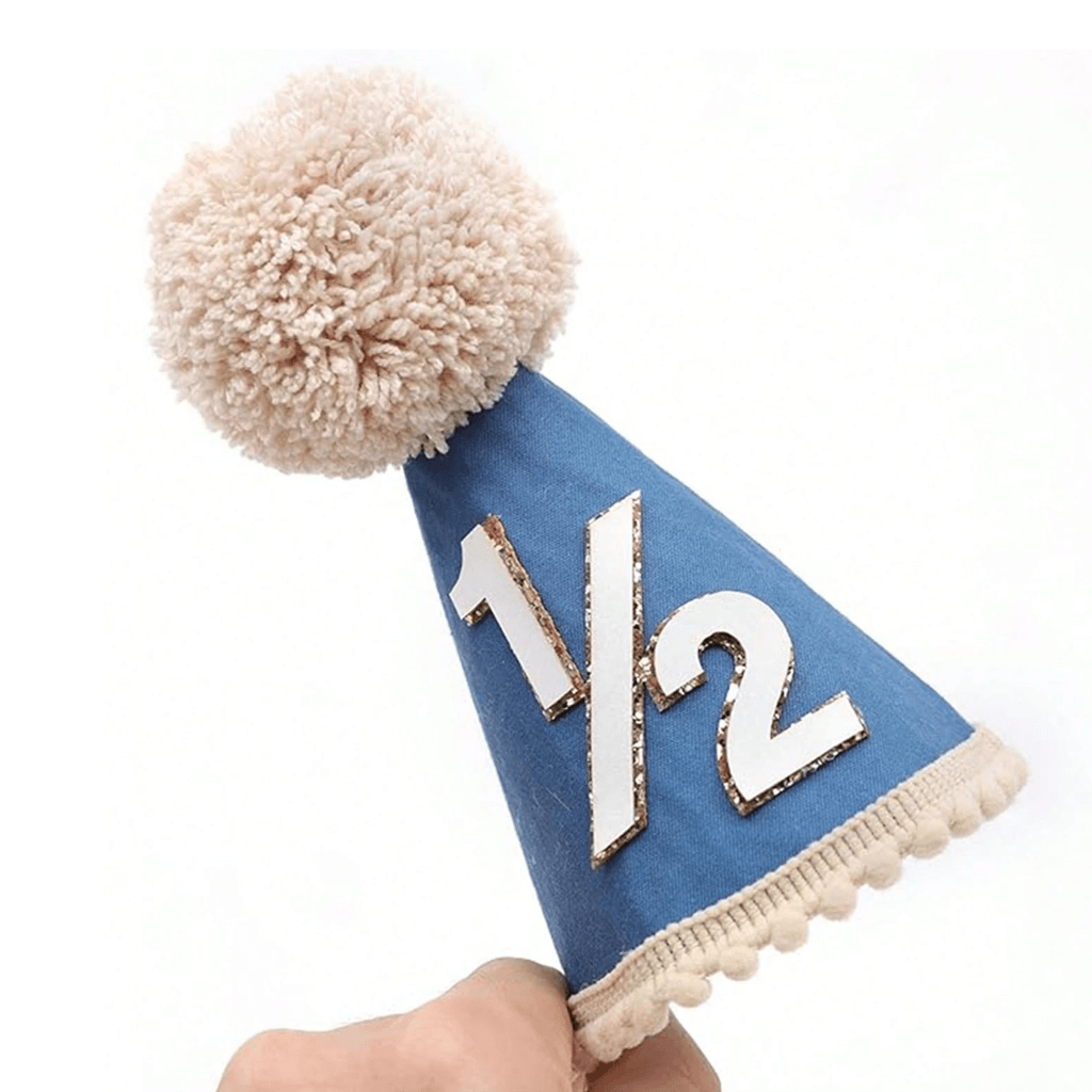 A blue baby half-year party hat with a beige pom-pom on top, white '1/2' text, and a beige ruffled trim, held by a hand against a white background.