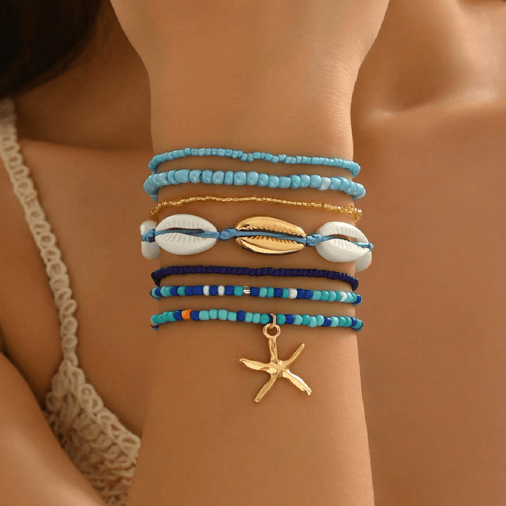 A stack of bohemian-style beaded bracelets on a woman's wrist, featuring light blue, navy, white, and gold beads, cowrie shells, and a gold starfish charm.