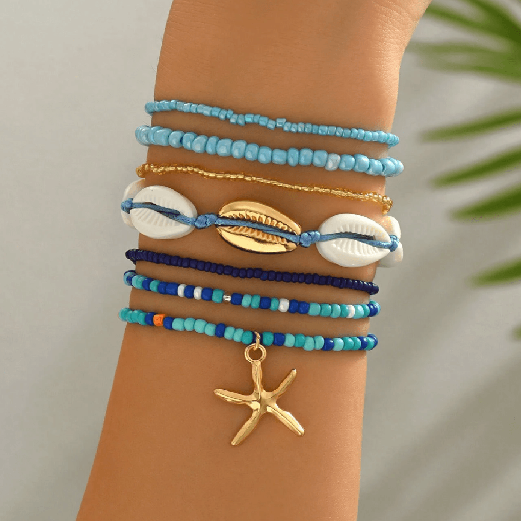 A stack of seven bohemian-style beaded bracelets on a person's wrist, featuring light blue, navy, and gold beads, white cowrie shells, a gold starfish charm, and a gold shell charm, with a soft-focus tropical background.