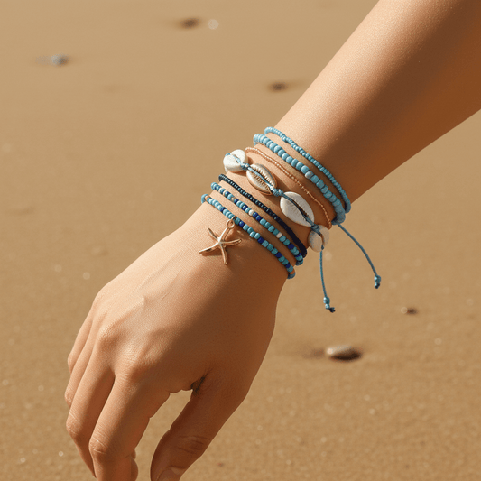 A close-up of a person's wrist and hand wearing a stack of beach-themed bracelets on a sandy beach. The bracelets include light blue beads, white cowrie shells, a small starfish charm, and a brown leather band.