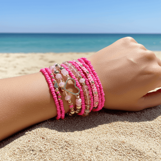Person's wrist with multiple pink beaded bracelets on a sandy beach.