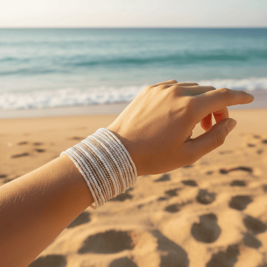 A close-up of a person's hand and wrist wearing a stack of multi-strand white beaded bracelets, set against a blurred beach background with sand and the ocean.