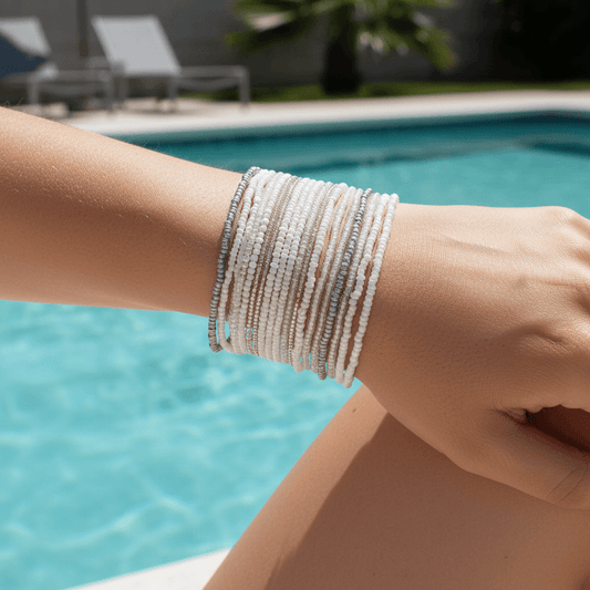 A close-up of a person's wrist wearing a stack of beaded bracelets in white, beige, and gray, with a blurred swimming pool in the background.