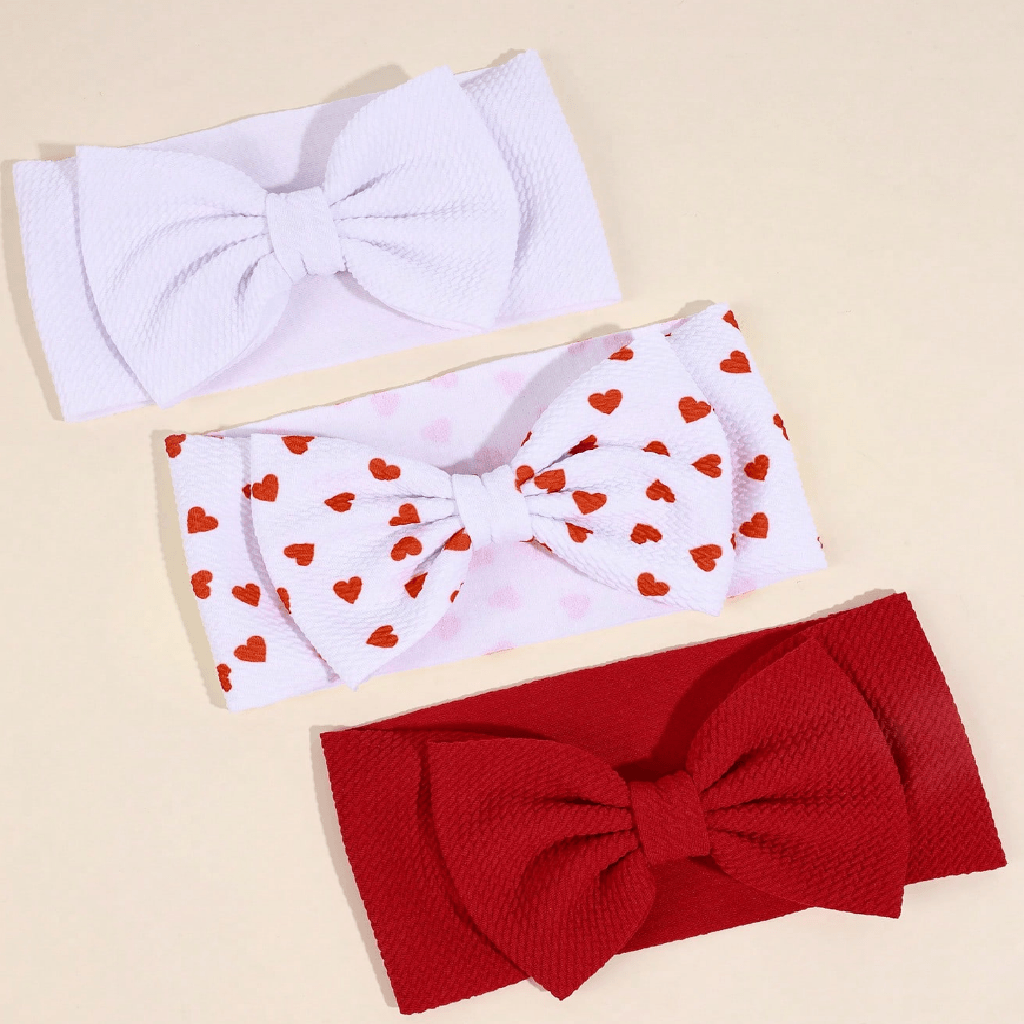 Three textured fabric bow headbands arranged diagonally on a light beige background: one solid white, one white with small red hearts, and one solid red.