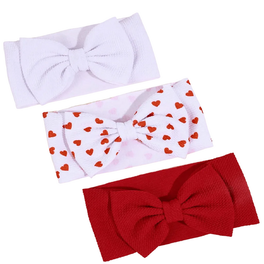 Three textured fabric bow headbands arranged diagonally on a light beige background: one solid white, one white with small red hearts, and one solid red.