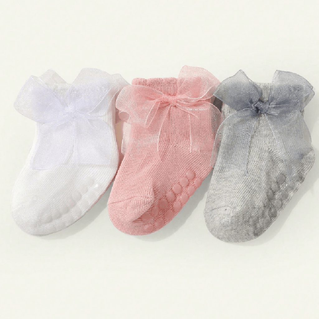 Three pairs of baby socks in white, pink, and gray, each featuring a sheer tulle bow at the top and textured grip soles, displayed on a light cream background.