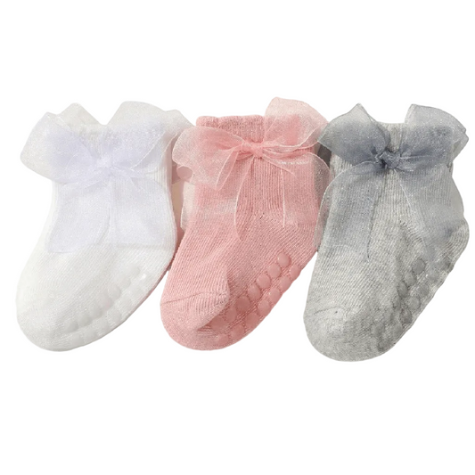 Three pairs of baby socks in white, pink, and gray, each featuring a sheer tulle bow at the top and textured grip soles, displayed on a light cream background.