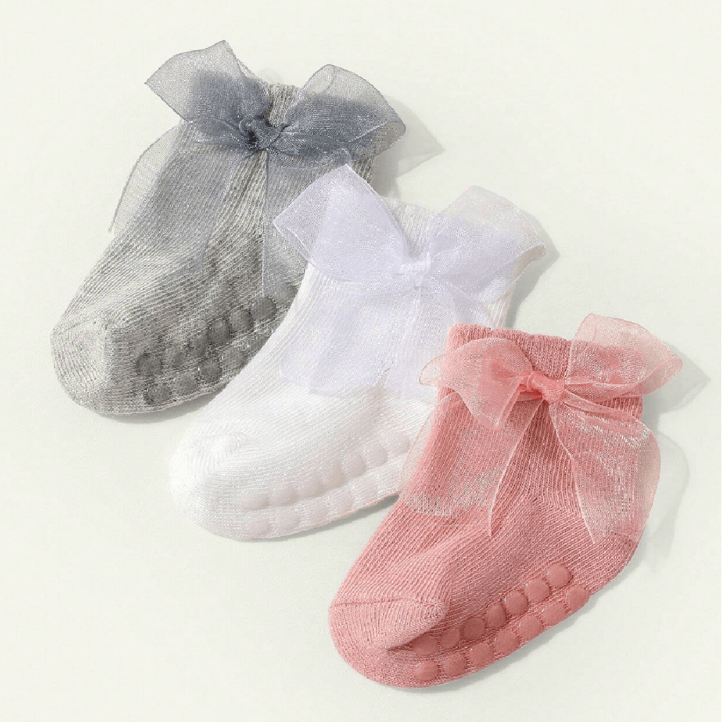 Three pairs of baby socks with sheer mesh bows and textured grip soles, shown in gray, white, and pink on a light cream background.