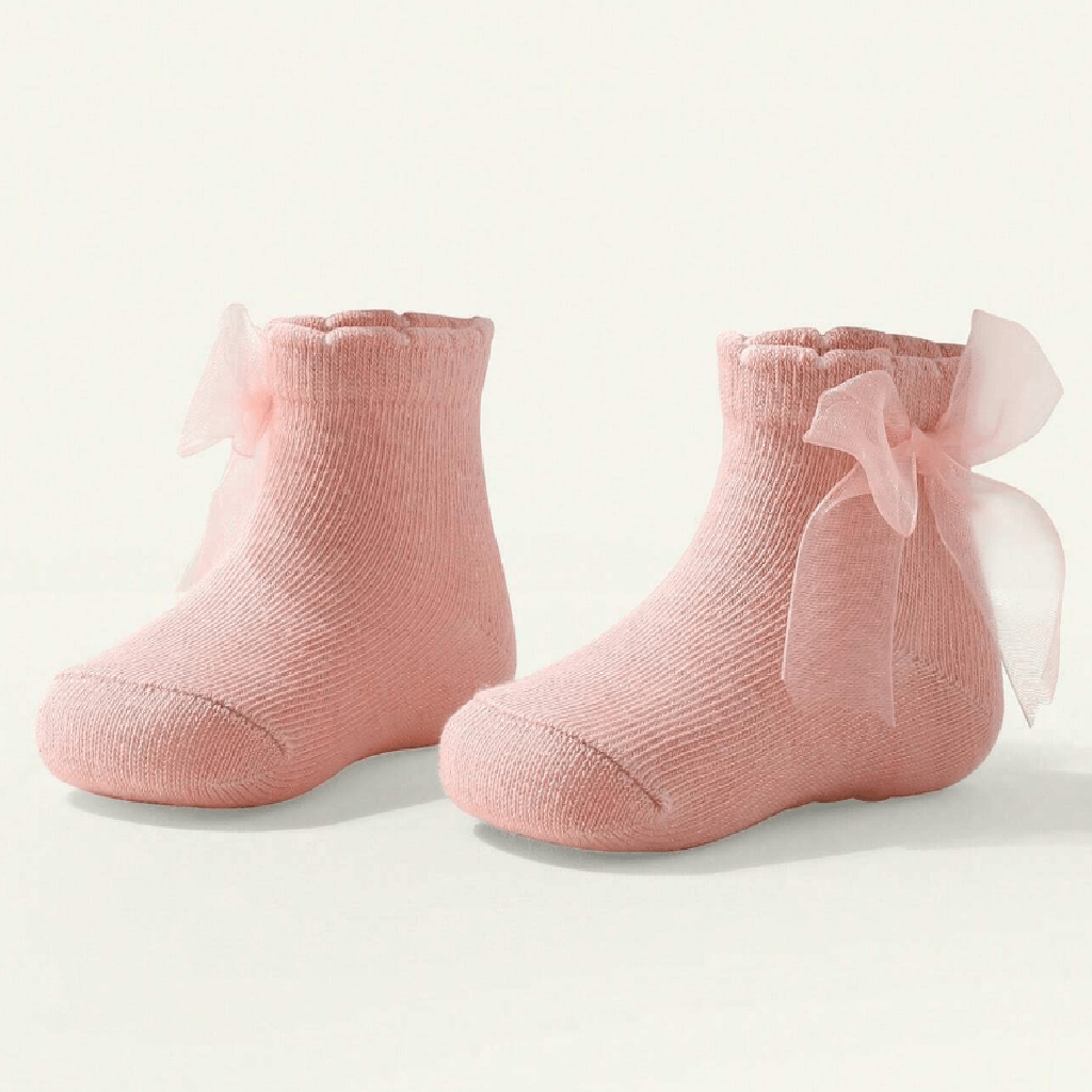A pair of soft pink knit baby booties with ruffled trim and large sheer pink ribbon bows, photographed on a clean off-white background.
