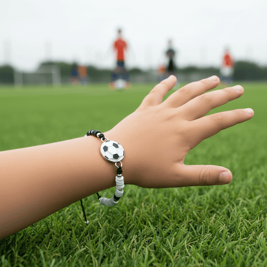 Child's hand wearing a soccer-themed bracelet on a grassy field with blurred figures in the background
