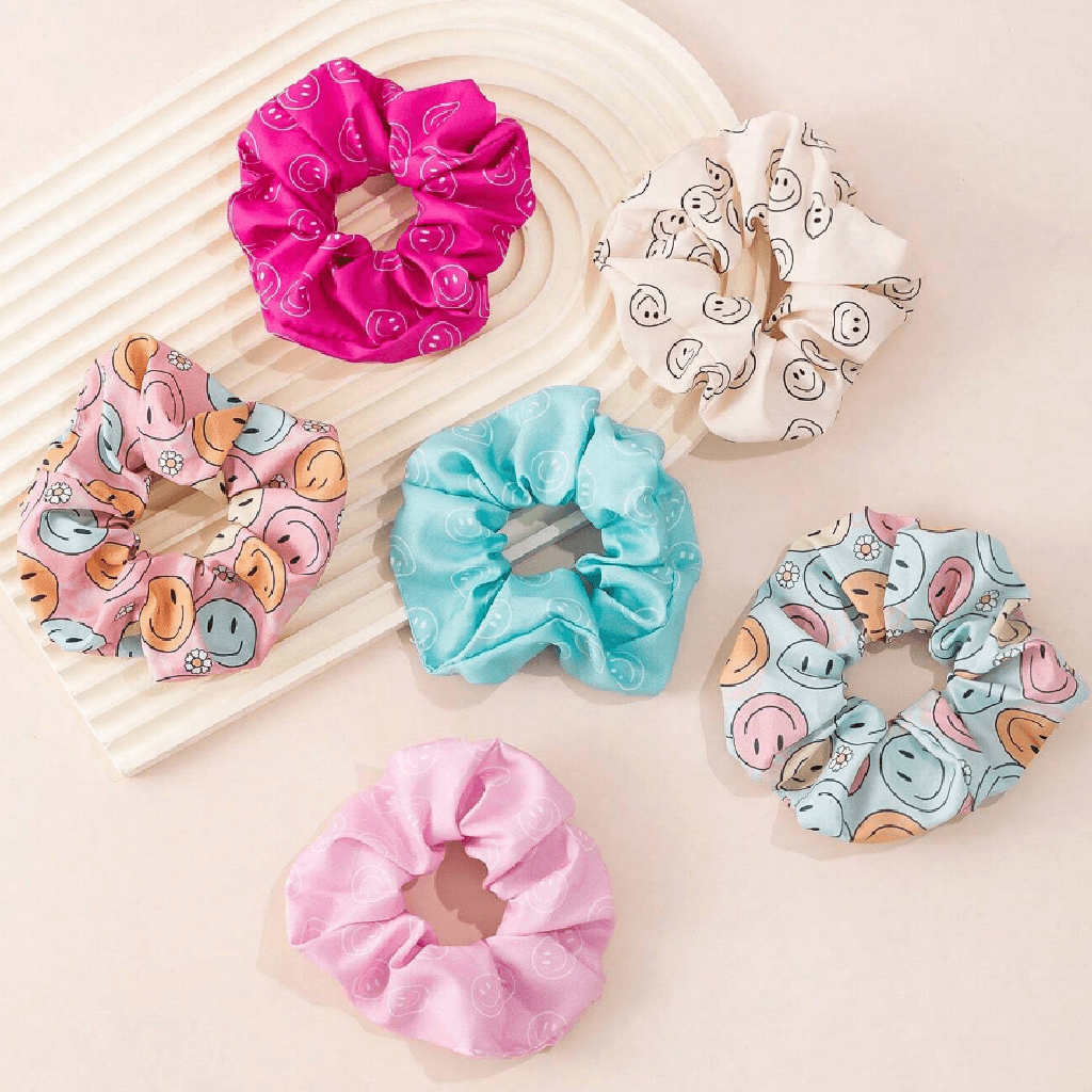Six fabric hair scrunchies in assorted colors and patterns, including smiley faces, donuts, and hearts, arranged on a light beige background with a white ribbed tray.