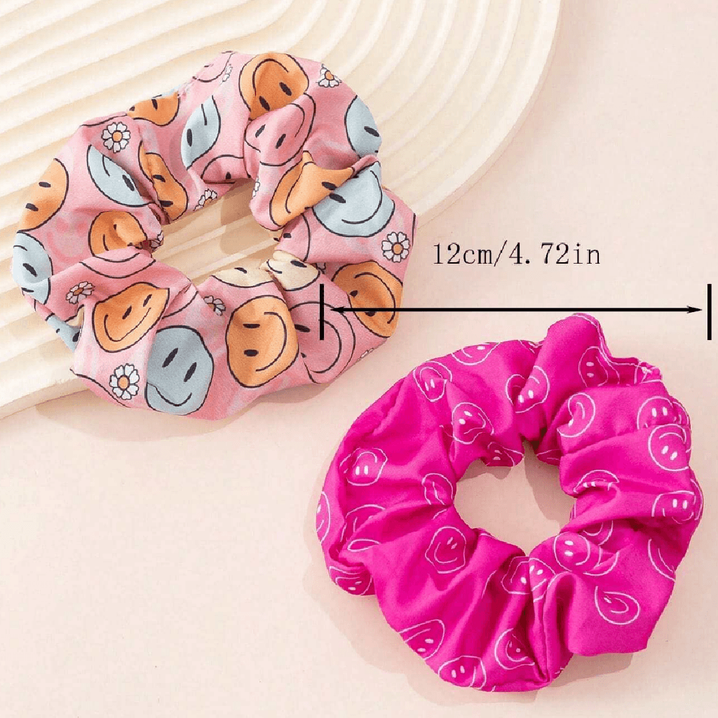 Two fabric hair scrunchies displayed on a light beige surface: one pink with a smiley face pattern and one bright pink with a white smiley pattern. A measurement arrow indicates the width of the larger scrunchie as 12cm / 4.