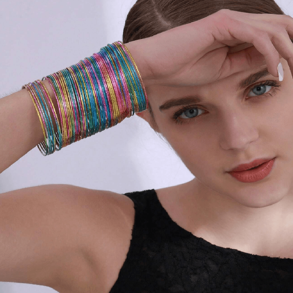 A close-up of a model wearing a stack of thin, iridescent bangle bracelets on her wrist. The bracelets shimmer with multiple colors including gold, pink, blue, green, and purple.