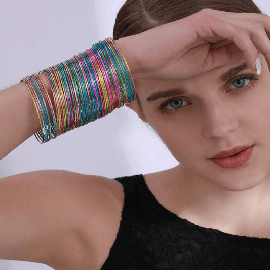 A close-up of a model wearing a stack of thin, iridescent bangle bracelets on her wrist. The bracelets shimmer with multiple colors including gold, pink, blue, green, and purple.