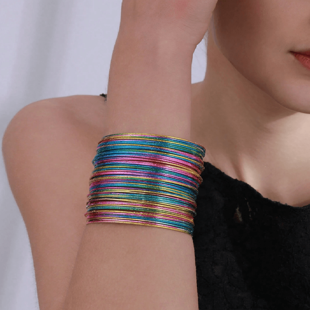 A close-up of a person's wrist wearing a stack of thin, iridescent metallic bangles in multiple colors including blue, green, pink, and gold.