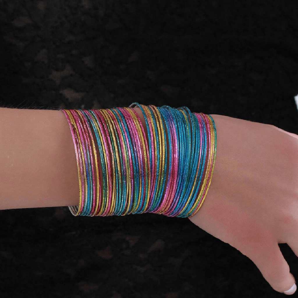 A close-up of a person's wrist wearing a stack of multiple thin, colorful metallic bangles or bracelets in pink, blue, green, yellow, and orange against a dark background.