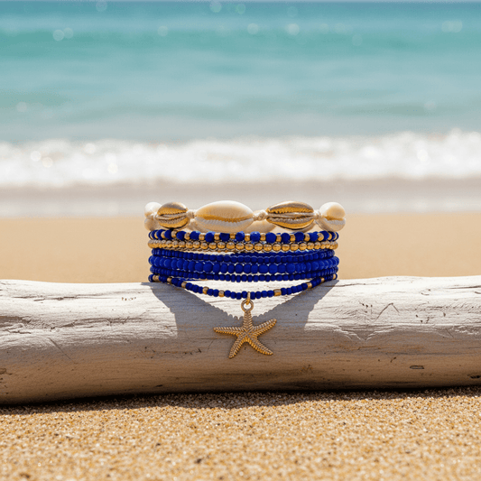 A stack of beaded bracelets with shell and starfish charms rests on a driftwood log on a sandy beach with the ocean in the background.