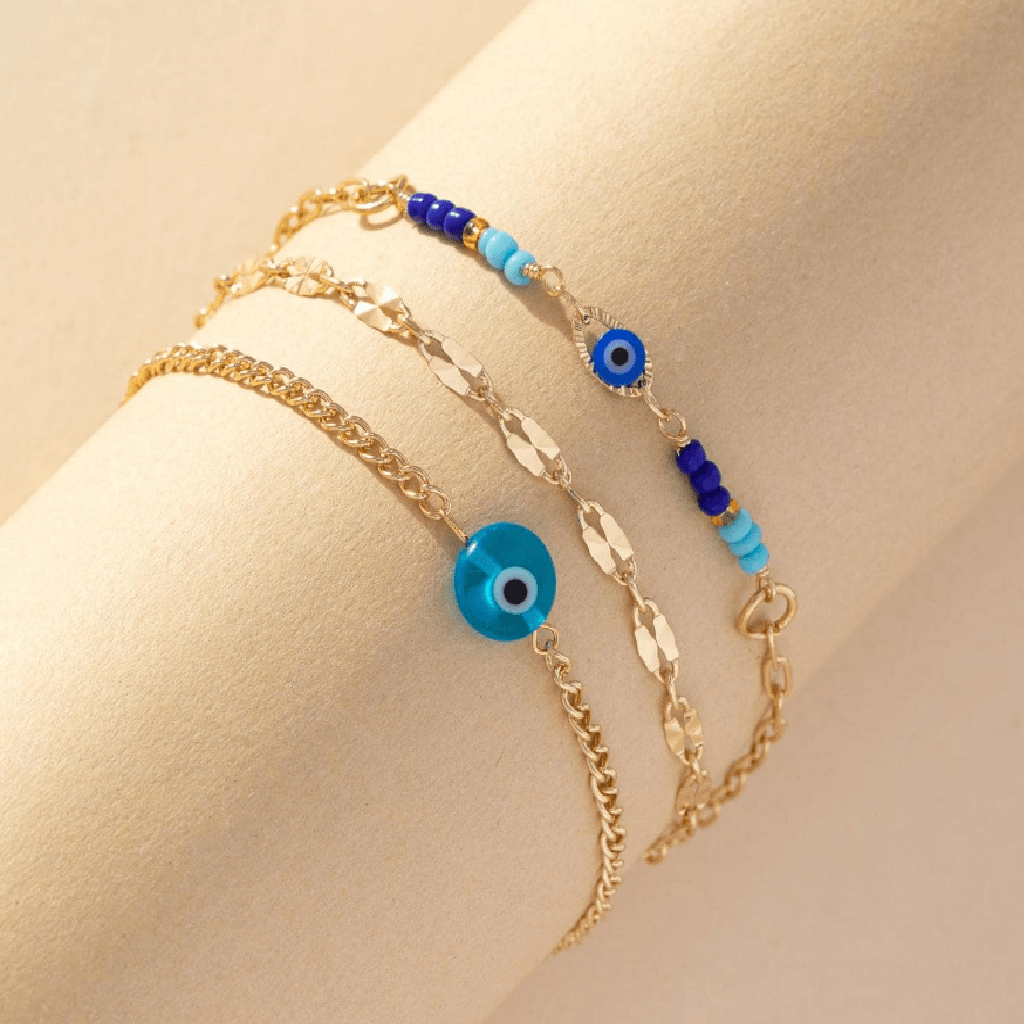 Three delicate gold-tone chain bracelets with evil eye bead details, displayed on a beige mannequin wrist. One bracelet features a large round blue evil eye charm, while the other two feature smaller blue and dark blue evil eye beads accented with light blue beads.