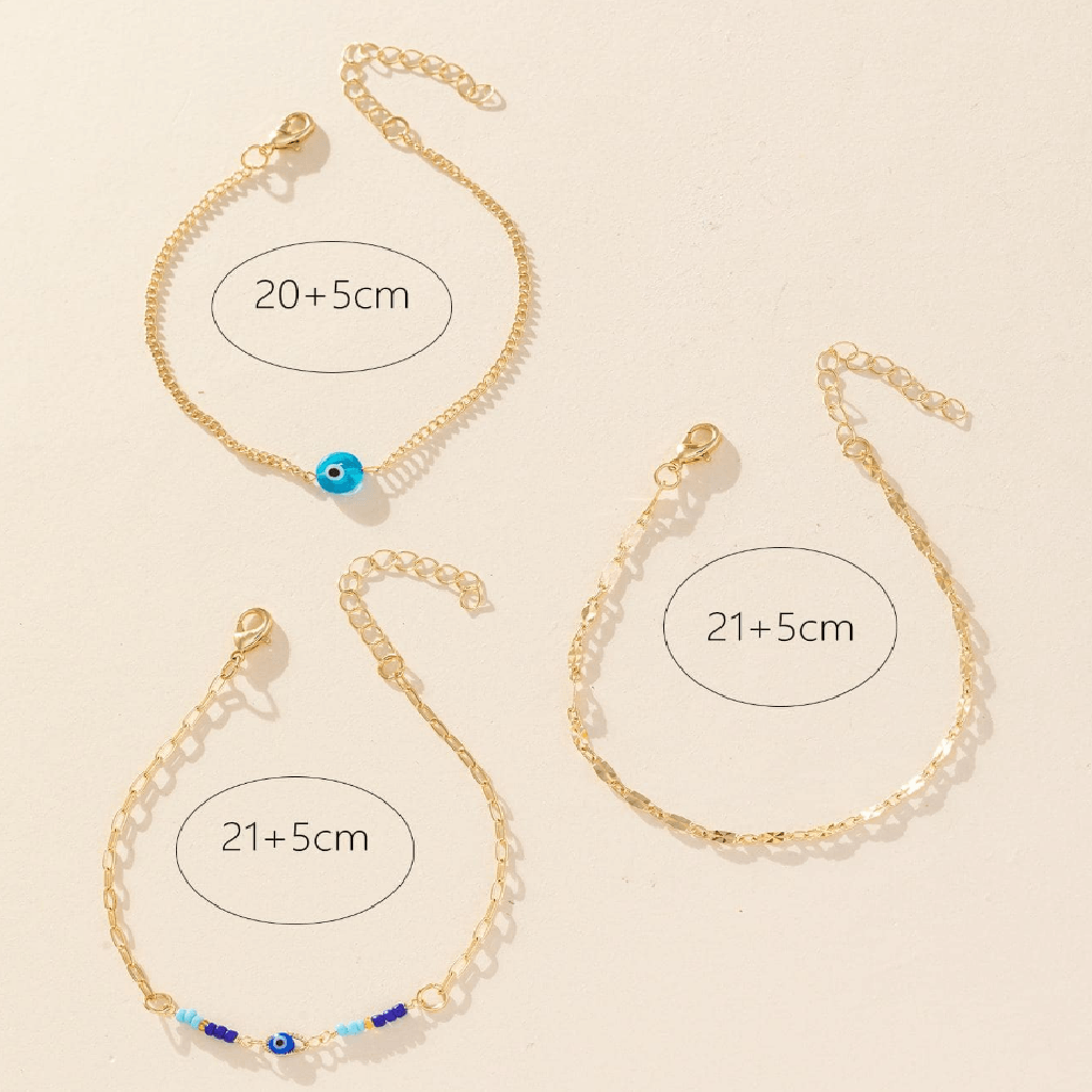 Three gold chain anklets with adjustable lobster clasp closures are arranged on a light beige background. One anklet has a small blue evil eye bead, and two anklets have small blue and white beads at the front.