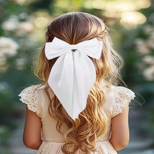 Girl with long blonde hair wearing a large white bow, standing outdoors with a blurred natural background.