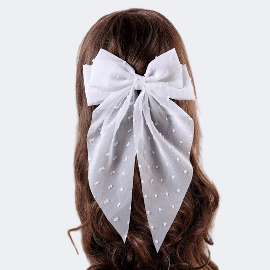 Woman with long blonde hair wearing a large white polka dot bow in her hair, against a plain background.