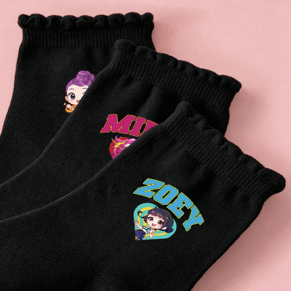 Three black crew socks with ruffled cuffs, featuring colorful cartoon character prints and the names 'MIRA' and 'ZOEY' on a soft pink background.