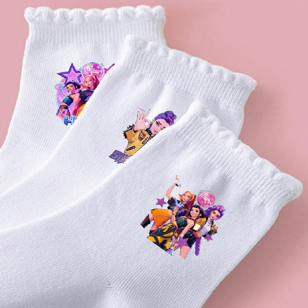 Three white crew socks with ruffled cuffs, each printed with a colorful cartoon character graphic, arranged diagonally on a soft pink background.