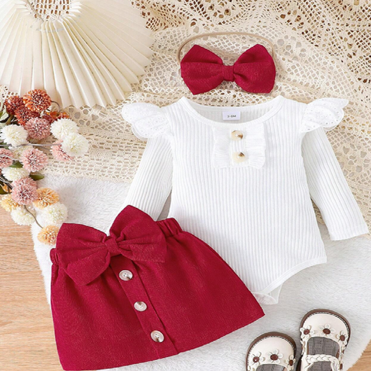 Baby outfit with white long-sleeve top and red skirt on a textured surface with flowers and a hat.