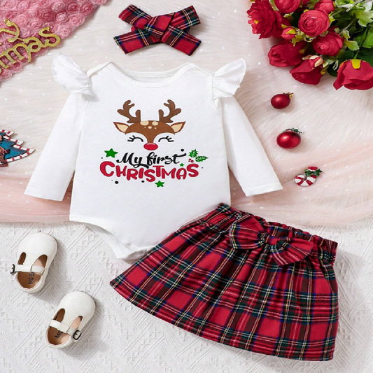Baby outfit with 'My first Christmas' shirt and red plaid skirt on a white surface with Christmas decorations.