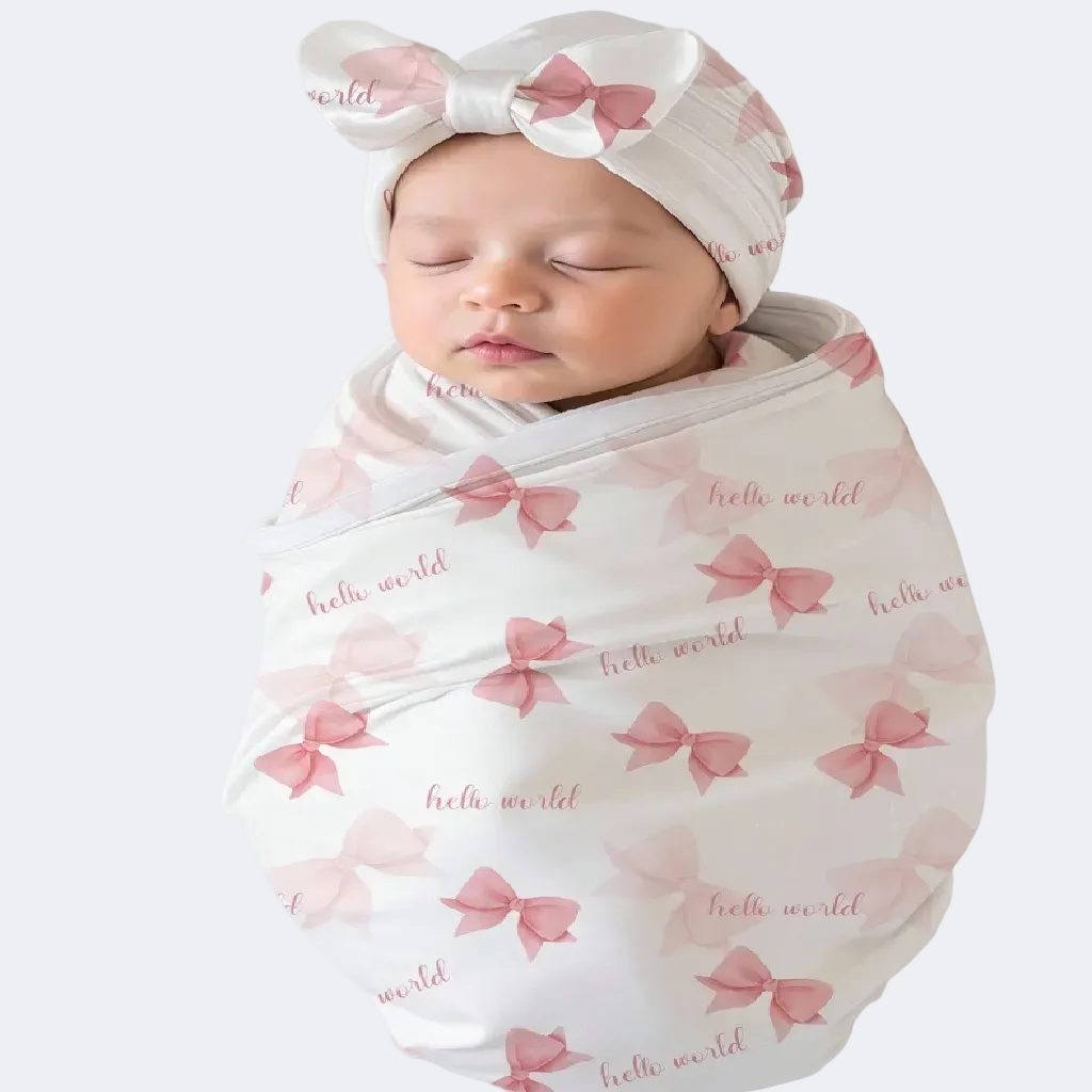 Newborn baby wrapped in a pink bow-patterned swaddle with 'hello world' text, wearing a matching headband.