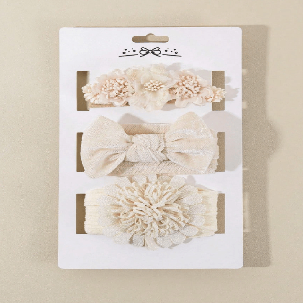 A three-piece baby hair accessory set in cream and beige, displayed on a white cardboard backing. The set includes a fabric flower headband, a knotted bow headband, and a large ruffled fabric flower headband.