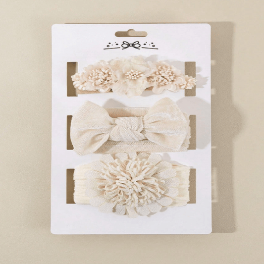 A three-piece baby hair accessory set in cream and beige, displayed on a white cardboard backing. The set includes a fabric flower headband, a knotted bow headband, and a large ruffled fabric flower headband.