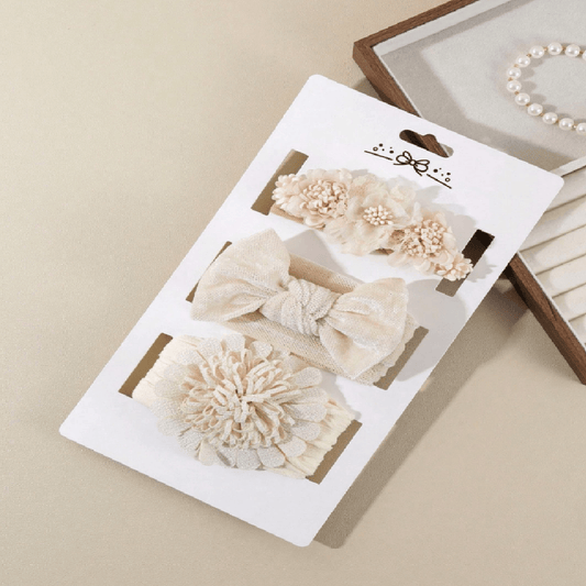 Three cream-colored hair accessories on a white display card, including two fabric bows and one textured flower piece, photographed on a beige surface with a jewelry box partially visible in the background.